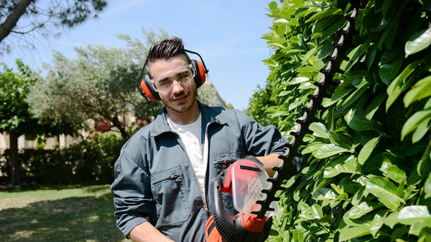 Védőszemüveget viselő fiatal férfi sövényvágó kerti szerszám használatával rendezi a sövénnyírást a kertben