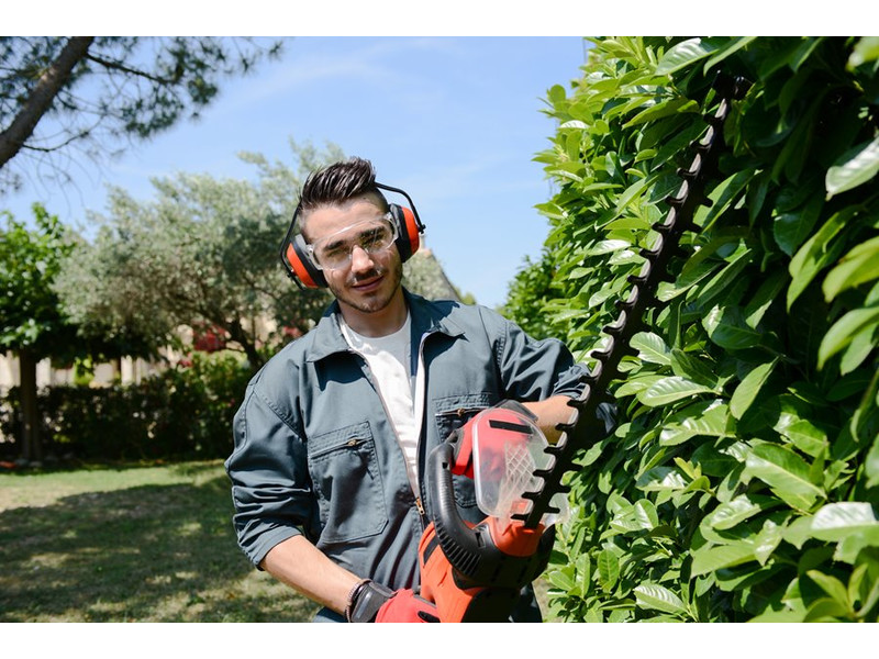 kert gépek és szerszámok a kertészkedéshez Védőszemüveget viselő fiatal férfi sövényvágó kerti szerszám használatával rendezi a sövénnyírást a kertben